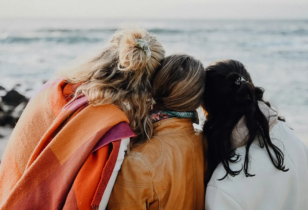 Three women seen from behind sit close together by the sea, heads leaning on one another, one wrapped in a blanket.