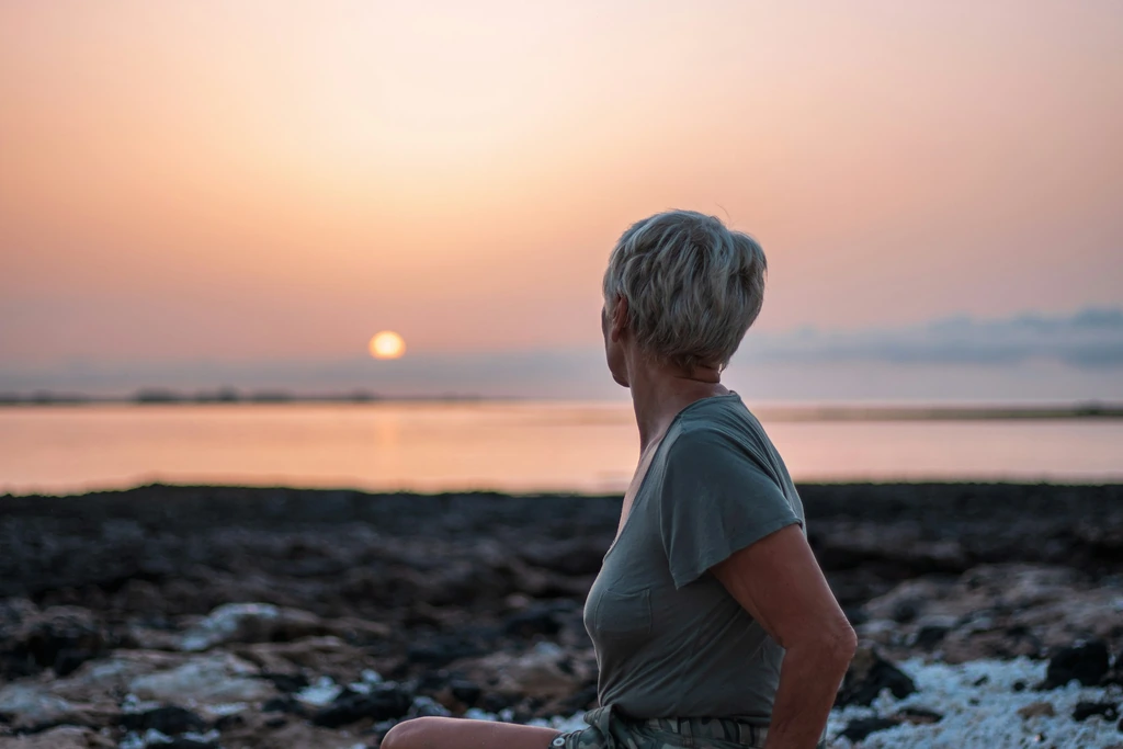 A woman with short grey hair, seen from behind, sits on a rocky shore watching a pink sunset over the water.